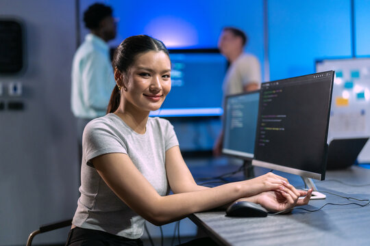 Young woman looks at camera and smiles while working at computer with code on monitor, female developer at workstation as colleagues work behind in blue lit office