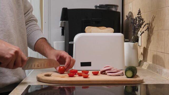 Person's hands chopping fresh cherry tomatoes on a wooden cutting board while preparing a nutritious breakfast or meal in a modern kitchen setting, emphasizing home cooking and food preparation