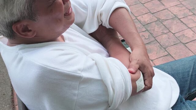 elderly asian man suffering from elbow tendonitis, having pain relieving patch on his elbow, clenching his jaw in pain on a park bench during daytime, high angle shot