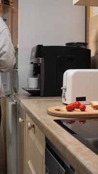 Vertical video man using a toaster to prepare breakfast with bread and fresh tomatoes on a cutting board in a home kitchen, creating a morning routine scene