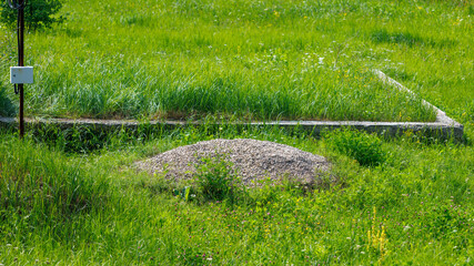 Naklejka premium Gravel mound sits in green grass. Concrete foundation lies nearby. Grass grows around construction site.