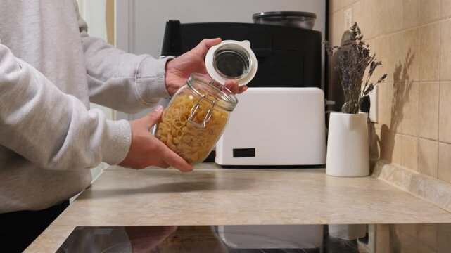 Person holding an open glass jar filled with uncooked shell pasta, preparing a meal in a domestic kitchen setting, emphasizing home cooking and food storage concepts