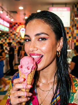 Young woman enjoying melting pink ice cream cone inside colorful ice cream parlor, smiling while licking treat with neon lights and busy interior in background