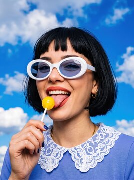 Woman with white round sunglasses holding yellow lollipop against blue sky with clouds, smiling outdoors in playful daytime portrait featuring retro lace collar styling