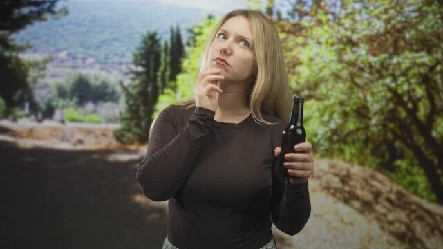 Woman holding beer bottle with right hand and touching chin while gazing upward on a forest path; thoughtful reflection.