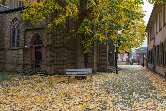 Golden autumn leaves surround a historic church and quiet bench in Zons, Germany