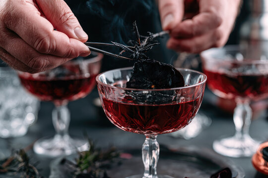 Adult hands using precision tweezers to place charred herb garnish on dark crimson cocktail in crystal glassware, creating dramatic Halloween serving scene with moody lighting and shallow focus
