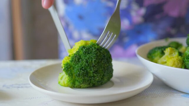 Steamed broccoli floret being cut with knife and fork on a white plate