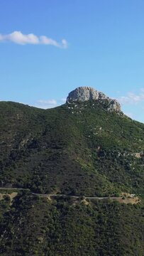 Vertical 9:16 aerial video showing a rugged, rocky mountain peak surrounded by lush green hills and a winding road below, captured between Baunei and Dorgali in Sardinia, Italy.
