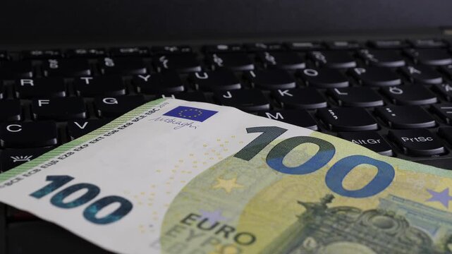 Close-up view of a 100 Euro banknote resting on a laptop keyboard, showcasing the intricate design of the currency and the keys of the device, emphasizing the connection between money and technology