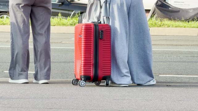 Travelers stand beside a bright red suitcase on a street in Saint Petersburg. They wait calmly for their transport to arrive. The bright luggage brings feelings of excitement and eager joy.