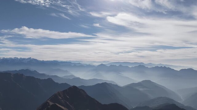 Layered dark mountain silhouettes recede toward glowing snow capped Himalayan summit under vast pale blue sky