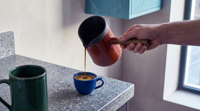Hand pouring coffee from a pot into a blue cup on a kitchen counter  