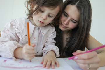 A little girl focuses on drawing with colored pencils while her mother sits by her side supporting her creative activity at a table at home. © atikinka2
