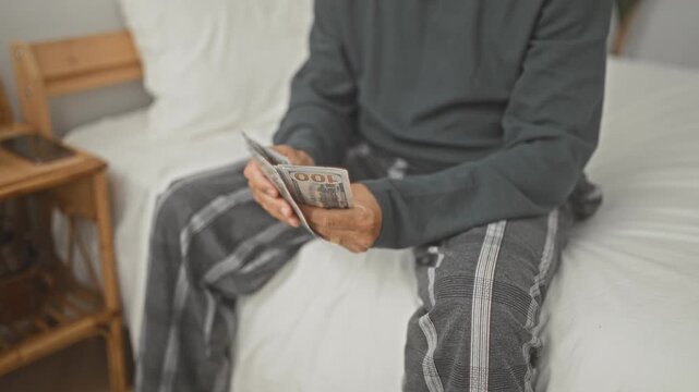 Man counting bills with hands on bed in a building, wearing pajama pants and a long sleeve top; anxiety savings.