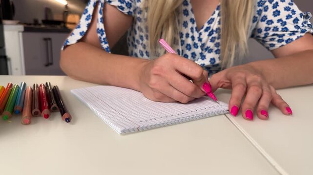 Close-up of hands writing with pink pen on squared notebook at white tabletop in home kitchen setting, with writing, hand, pen, notebook detail, showing pencil, colored, nail, pink context