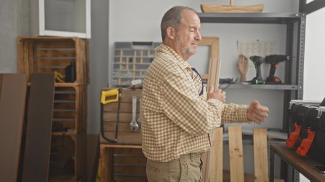 Man carpenter with arms crossed holding wood plank in building with drills and toolbox on shelves; craft pride.
