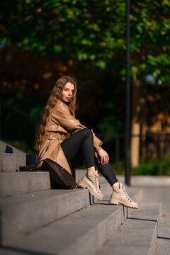 woman relaxing on steps in casual outfit in urban park setting with brown leather jacket and beige ankle boots, side view slightly elevated angle, casual and relaxed mood.