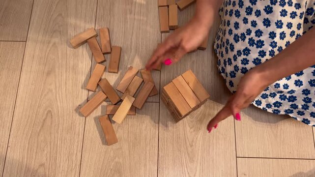 Low-angle interior motion scene of hands manipulating numbered wooden blocks on light wood-look laminate floor, with hand, block, tower, stack detail, showing number, wood, wooden, floor context