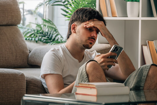 Stressed young man sitting on sofa at home looking worried at smartphone screen, frustrated student feeling anxious and overwhelmed while reading bad news or message, mental health and stress concept