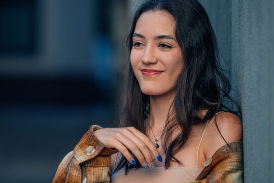 smiling young woman with long dark hair leaning against a wall, looking away thoughtfully with a warm autumn light