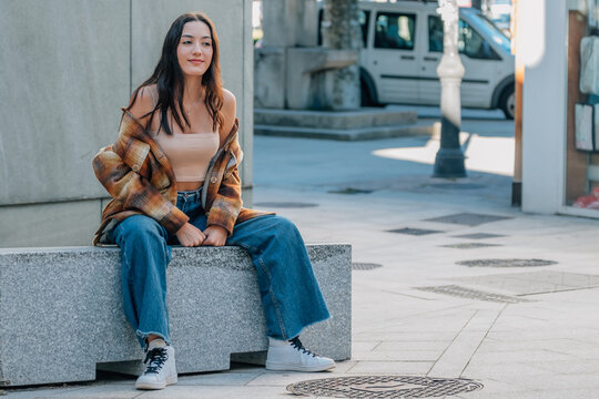 Young woman with long brown hair sitting outdoors on a stone bench, smiling and looking away, wearing a plaid flannel shirt jacket, autumn urban lifestyle portrait