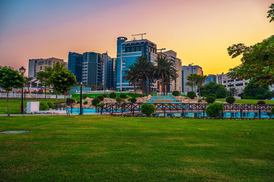 Colorful sunset over Al Mathaf Park in Doha, Qatar, with green lawns, palms, footbridges, water features, and a modern city skyline.