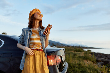 Fototapeta premium Woman with hat leaning against a car by the coast, wearing a striped shirt, denim jacket and yellow skirt, holding a phone. Sunny outdoor lifestyle moment.