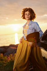Naklejka premium A woman with curly hair stands beside a car at sunset, wearing a striped shirt and denim jacket, capturing a relaxed lifestyle moment. Concept: casual confidence and outdoor freedom.
