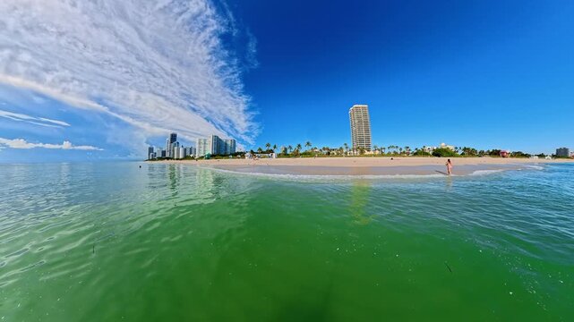 Camera approaches a sandy Miami Beach shoreline from clear green ocean water.
