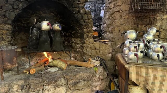 Slow forward camera movement toward a rustic wood-fired tea-making setup in a Turkish village. Multiple metal kettles are heated over an open fire on tall traditional metal vessels inside an old stone