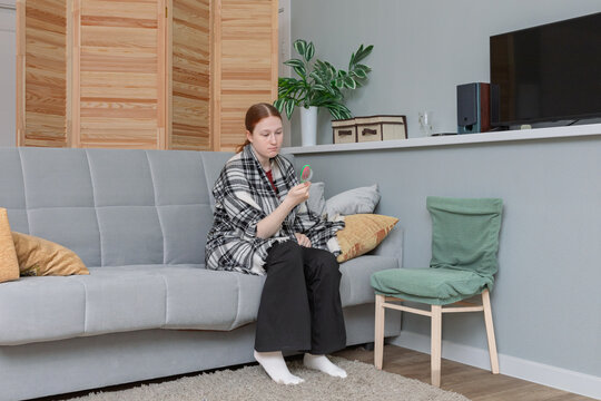 Young woman with red hair sitting on a sofa wrapped in a blanket and holding a small hairbrush at home