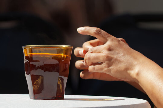 A hand reaches for a glass of chilled amber drink on a sunlit white table, warm sunlight and a blurred dark background suggest a relaxed cafe terrace or outdoor market moment.
