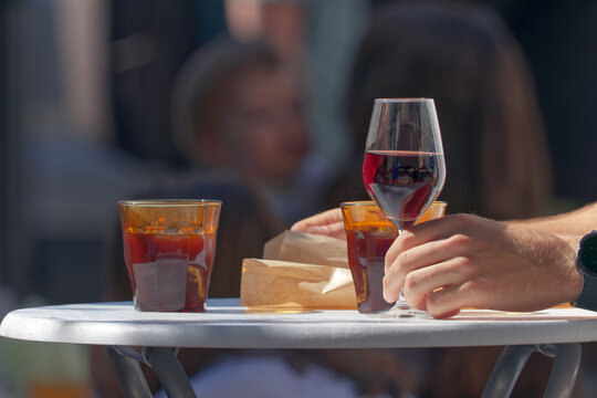 Red wine, amber glasses, and a paper food bag rest on a table at the farmers market Hermanak in Prague. The blurred background of socializing people highlights a vibrant community weekend lifestyle.