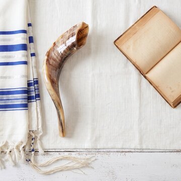 Flat lay of Jewish religious items for Rosh Hashanah and Yom Kippur with copy space. Shofar ram horn, tallit prayer shawl with blue stripes, and old Hebrew prayer book on white background. 