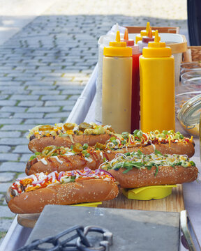 Five loaded gourmet hot dogs with fresh toppings sit before condiment bottles on a cobblestone street at the Naplavka farmers market in Prague.
