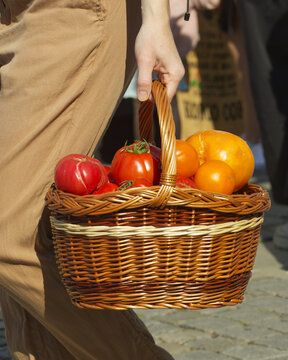 Close-up vertical shot of a hand carrying a woven basket filled with red and orange heirloom tomatoes at farmers market Naplavka in Prague, highlighting fresh seasonal produce.