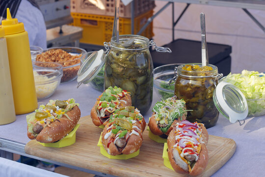 Five loaded gourmet hot dogs with colorful toppings rest on a wooden board beside pickle jars and condiment bottles at a Naplavka farmers market stall in Prague.
