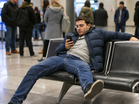 Traveler sprawls across several terminal seats while using a phone, reducing seating for others and illustrating poor etiquette in a shared travel space.
