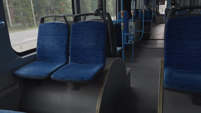 The interior of a public bus features blue upholstered seats arranged in rows, with a clear aisle leading to the front. Natural light enters through the windows, illuminating the bus's interior space