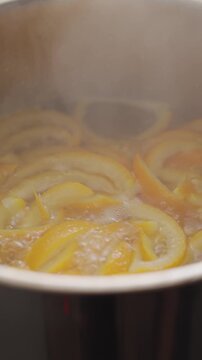Candied orange peel boils in syrup inside saucepan, with steam rising over the citrus strips. Concept of homemade orange candy and dessert recipe