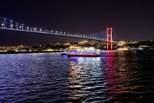 A nighttime Bosphorus Dinner Cruise boat sails on the Bosphorus Strait with the colorfully illuminated Bosphorus 15 July Martyrs Bridge behind it connecting Asia and Europe, in Istanbul, Turkey.