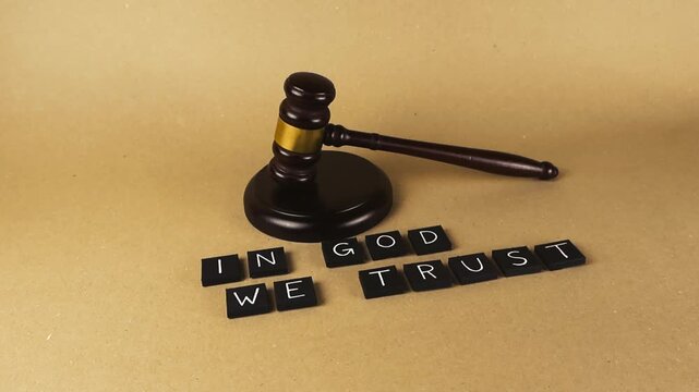 Smooth camera movement showing a wooden judge gavel resting next to the motto In God We Trust spelled out with letter tiles on a brown background, representing law, faith, and justice