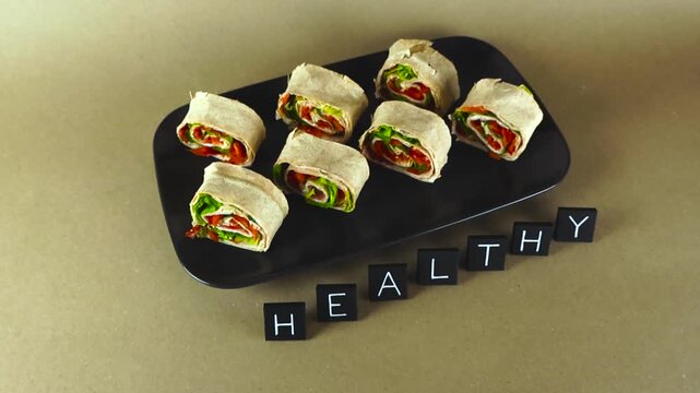 Fresh lavash pinwheel rolls with ham and vegetables served on a black plate with the word healthy spelled out in tiles, demonstrating a nutritious meal concept on a brown background