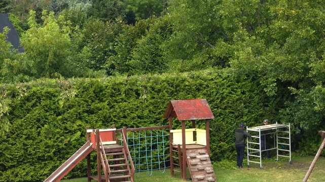Two professional gardeners collaborating to prune and shape a large, overgrown hedge in a backyard with a playground, one climbing a scaffold to use a powerful hedge trimmer for landscaping