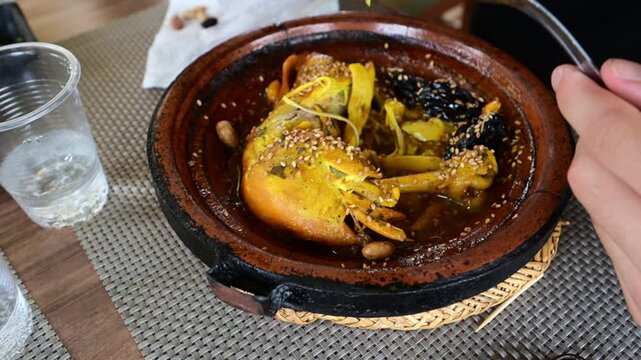 Marrakech, morocco. August 8, 2026. Person enjoying a delicious moroccan chicken tagine with preserved lemons, prunes, and sesame seeds served in a traditional clay pot