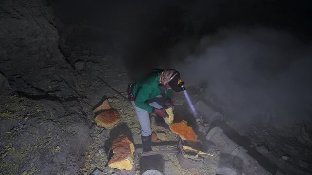 Local miner collecting and loading raw sulfur inside Kawah Ijen crater at night in East Java, Indonesia, surrounded by volcanic fumes and protected by masked ventilation, vertical