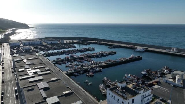 Wide aerial view of Tangier fishing port in Morocco, with crowded fishing piers, a protected harbor basin, long breakwater, coastal road and open sea.