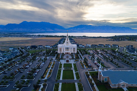 Saratoga Springs, Utah, United States - 22 August 2026: Aerial view of the Saratoga Springs Utah Temple glowing at blue hour, with surrounding lawns, parking lots, and residential neighborhoods.