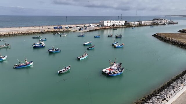 Wide aerial view of small fishing boats spread across the protected harbor of Asilah, Morocco, highlighting artisanal fisheries, calm coastal waters and working port infrastructure on the Atlantic coa